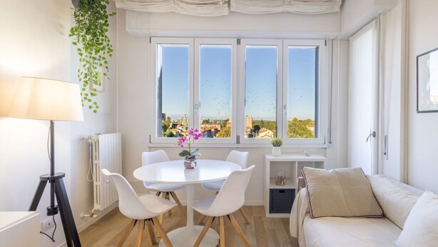 Une pièce lumineuse et moderne, avec une table de salle à manger ronde, des chaises blanches, un canapé et une grande fenêtre donnant sur la ville.