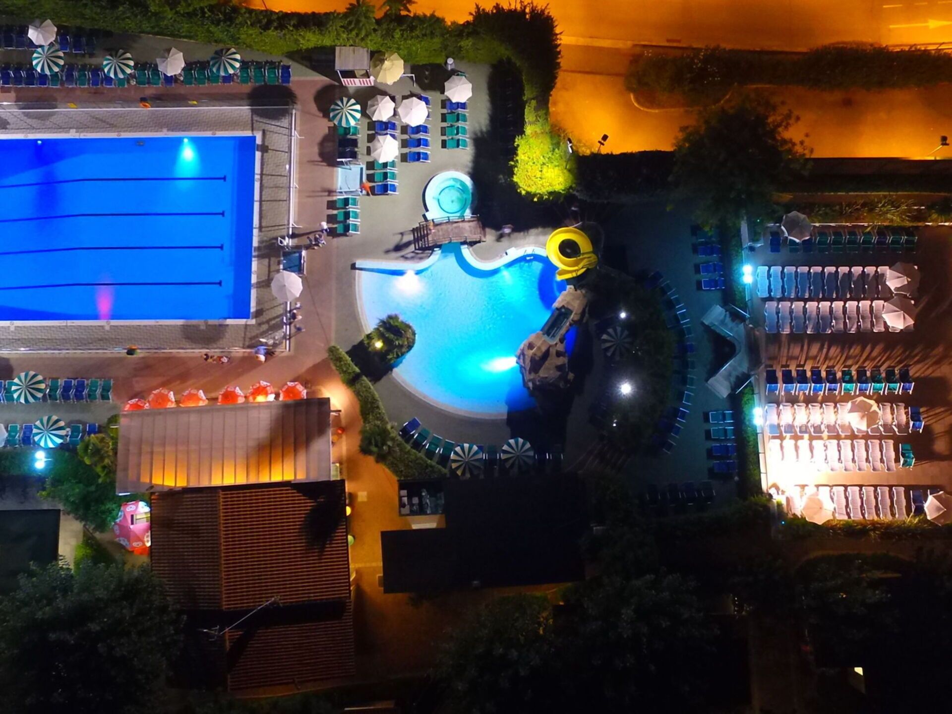 Eine Vogelperspektive zeigt ein Resort mit Swimmingpool, Liegestühlen, Sonnenschirmen und einer Wasserrutsche.