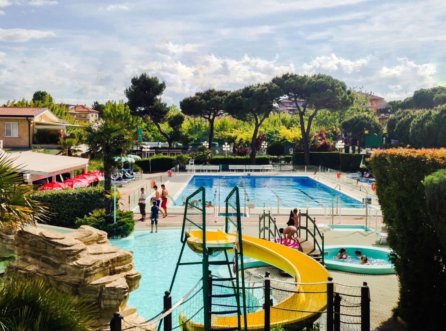 A swimming pool with a slide, surrounded by greenery and a few buildings in the background.