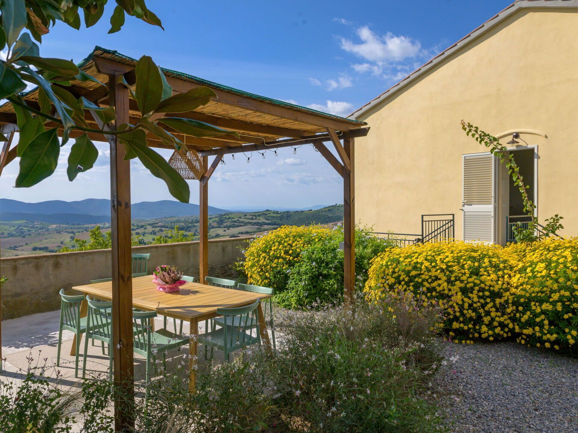 Un patio con una mesa y sillas de madera, rodeado de arbustos florecientes amarillos y vistas a colinas distantes.