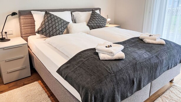 Ein Doppelbett mit grauem Kopfteil, weißer Bettwäsche und zwei Kissen. Ein Nachttisch mit Lampe, einer Schublade und einem gefalteten Handtuch befindet sich auf dem Bett.