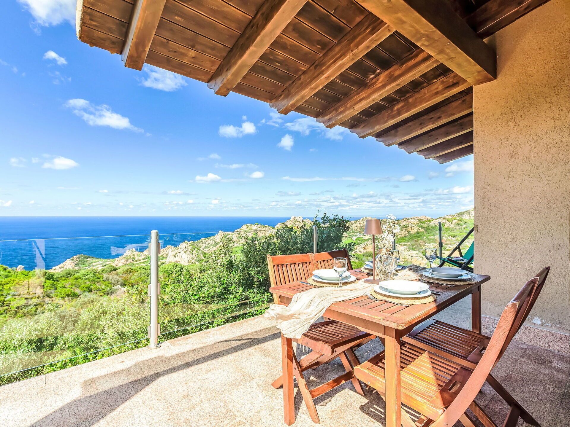 Ein Holztisch mit Stühlen steht auf einem Balkon mit Blick auf eine Küstenlandschaft, wo sich das klare Meer und ferne Hügel erstrecken.