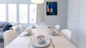 A modern dining area with a white table set for four, a painting on the wall, and a view of buildings through large windows.