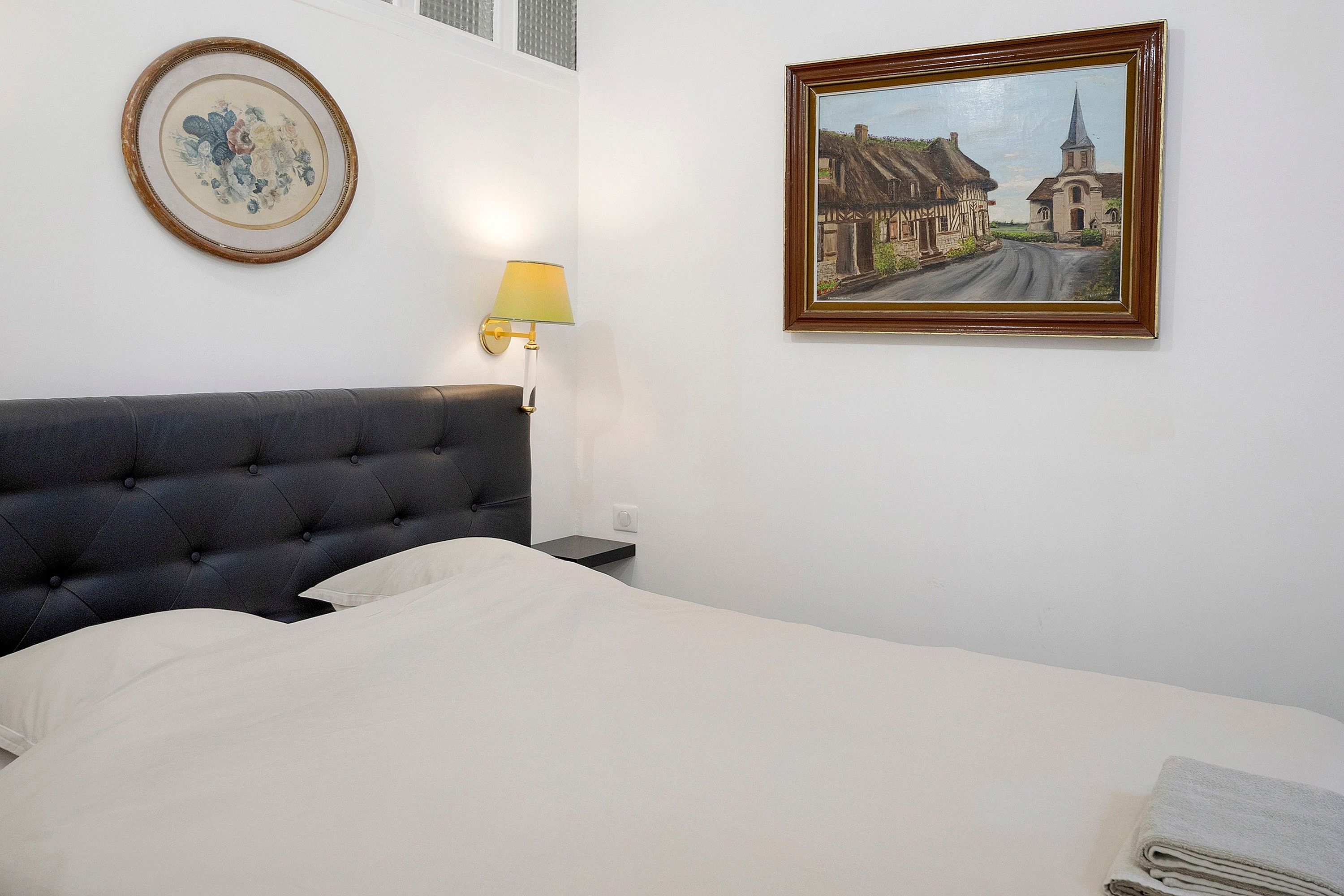 A hotel room with a bed, a framed painting, a lamp, and a wall-mounted picture frame.