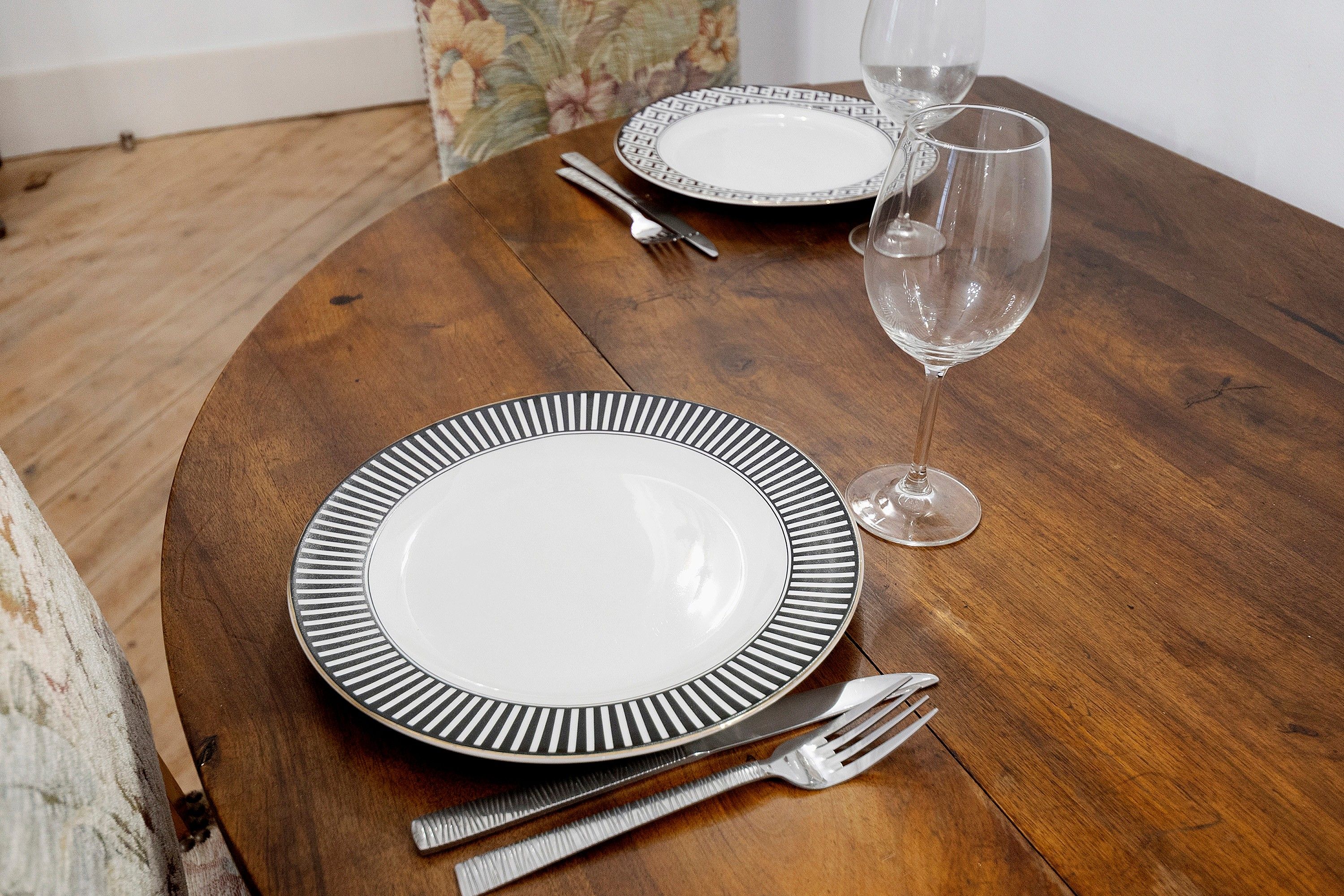 Een ronde houten tafel met een wit bord met een zwarte rand, een vork, een mes en een wijnglas.