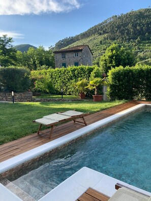 A pool area with a wooden deck and lounge chairs, surrounded by lush greenery and a stone building in the background. - Casa Locci, villa with swimming pool a stone's throw from Lucca (Capannori)