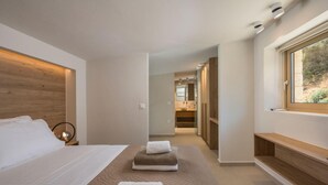 A modern hotel room with a large bed, a wooden headboard, a bedside table with a lamp, a bench by the window, and a view of the bathroom. - La Divina Villa (Zoe) (Chania)