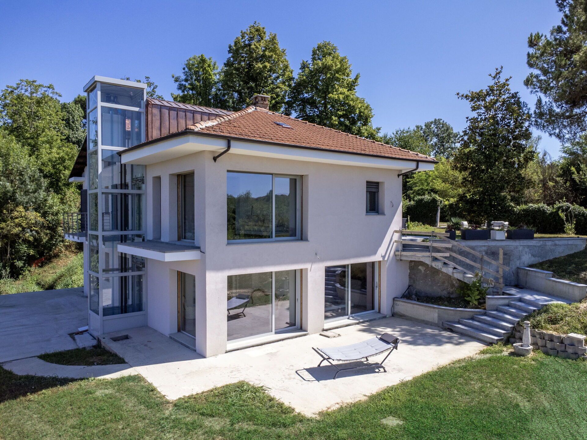 A modern two-story house with a balcony, large windows, and a patio with a sun lounger.