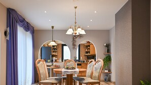 A dining area with a round table, chairs, and a chandelier. There is a kitchen visible through an archway.
