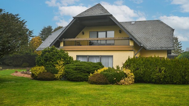 A two-story house with a sloped roof, a balcony on the upper floor, and a well-maintained lawn.