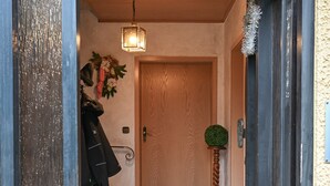 A wooden door with a doormat, a hanging coat, a potted plant, and a light fixture.