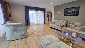 A living room with a sofa, armchair, and coffee table, a painting on the wall, and a view into the kitchen.