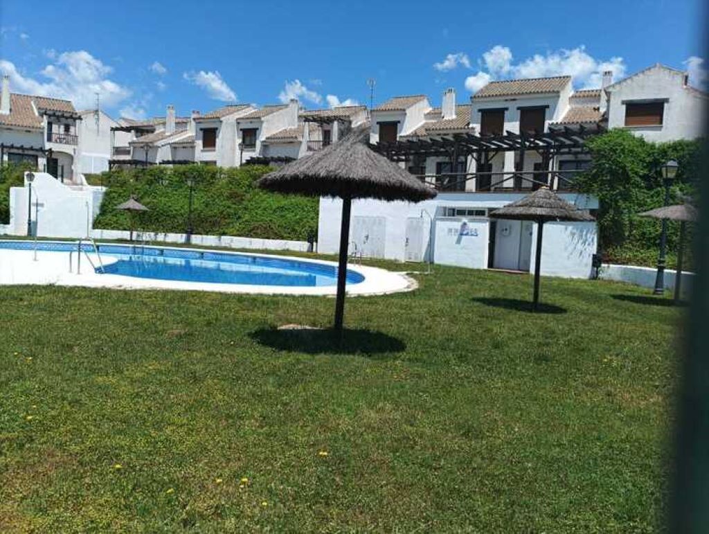 Une piscine agrémentée d’un parasol en chaume, entourée d’une pelouse et de bâtiments blancs aux toits de tuiles.
