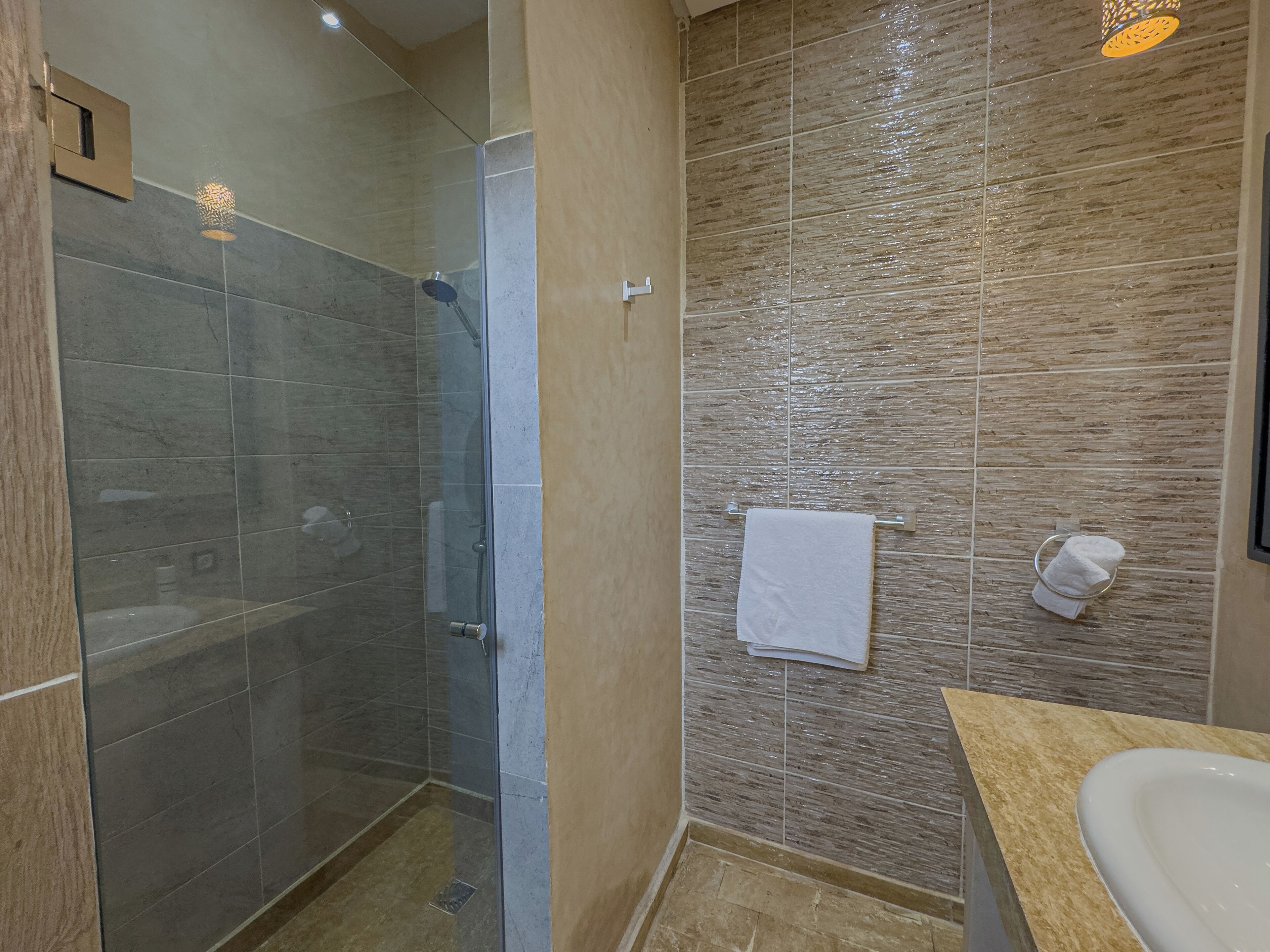 A modern bathroom with a glass-enclosed shower, a beige tiled wall, a white sink, and a towel rack.