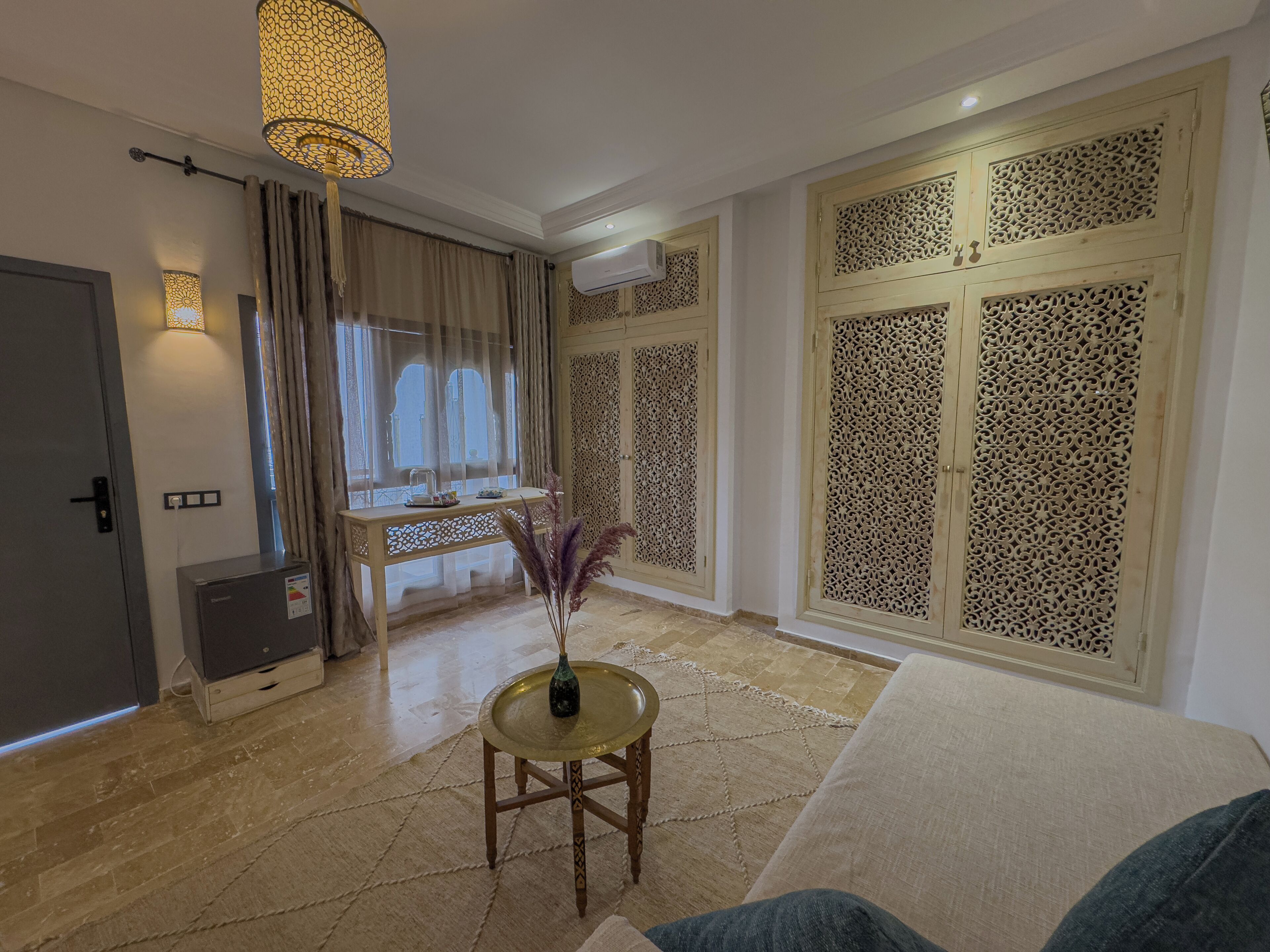 Een woonkamer met een bank, een klein rond tafeltje met een vaas, een raam met gordijnen en een decoratief roomdivider.