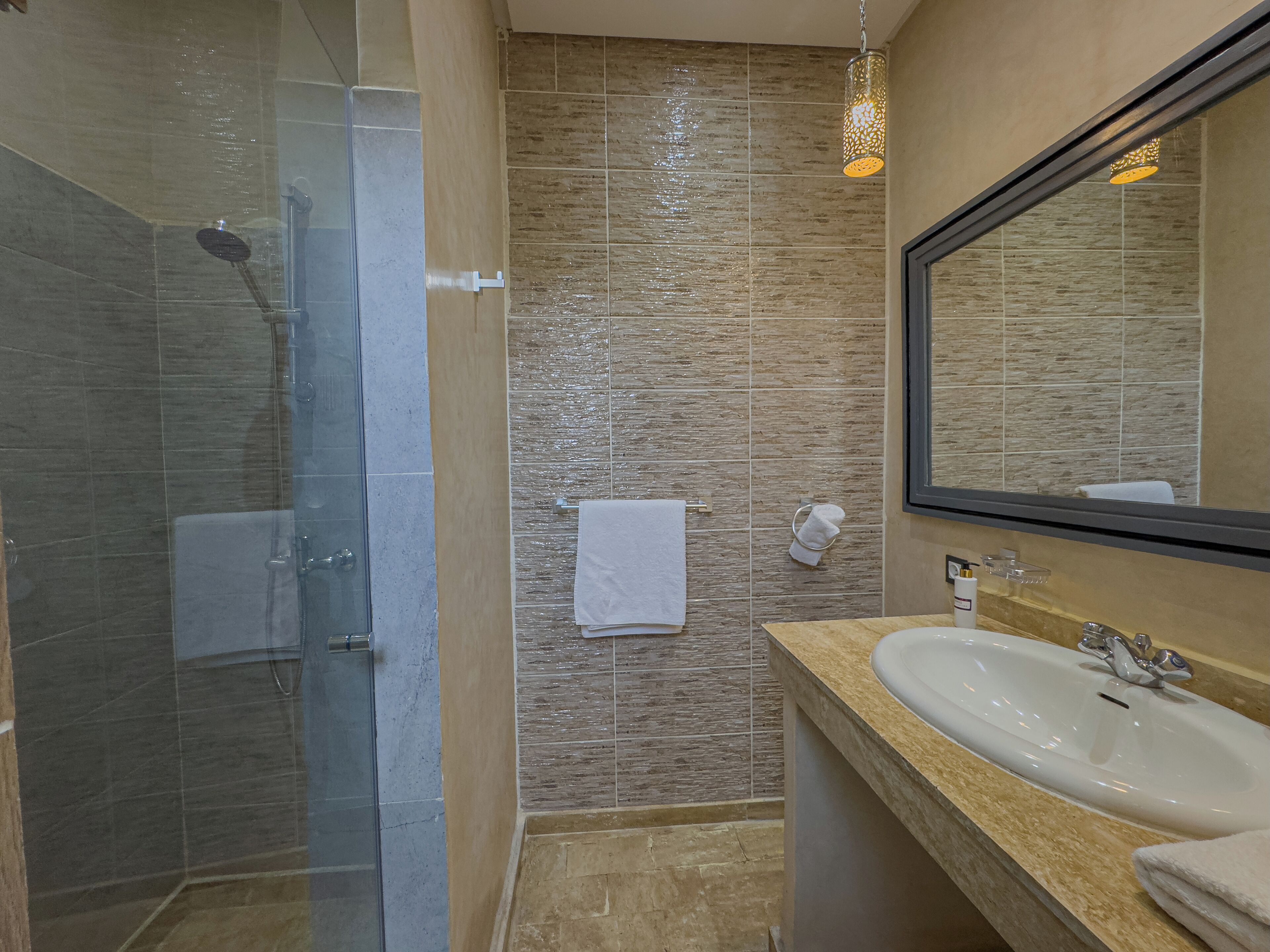 Un baño moderno con ducha de vidrio, pared con azulejos color beige, lavamanos blanco y un espejo grande.