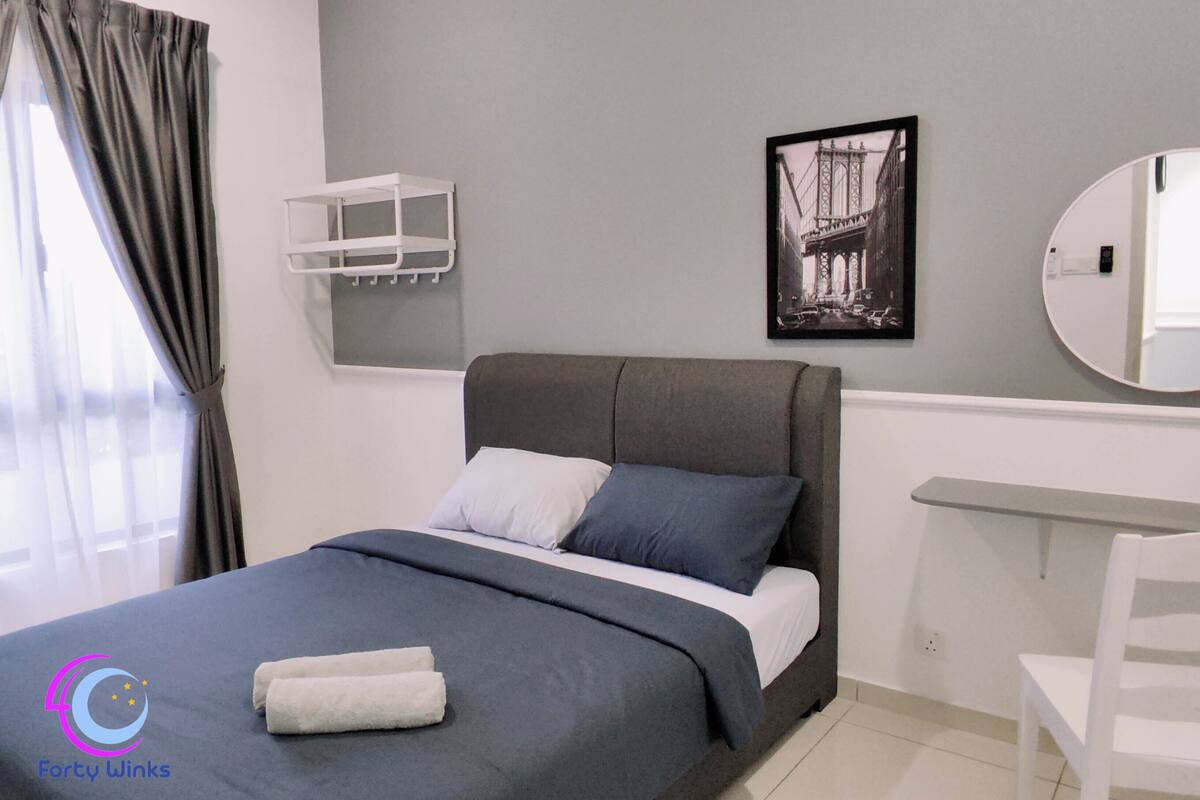 A modern bedroom with a grey bed, white and blue pillows, a grey headboard, a white bedside table, a round mirror, a framed picture of a bridge, and a white towel on the bed.
