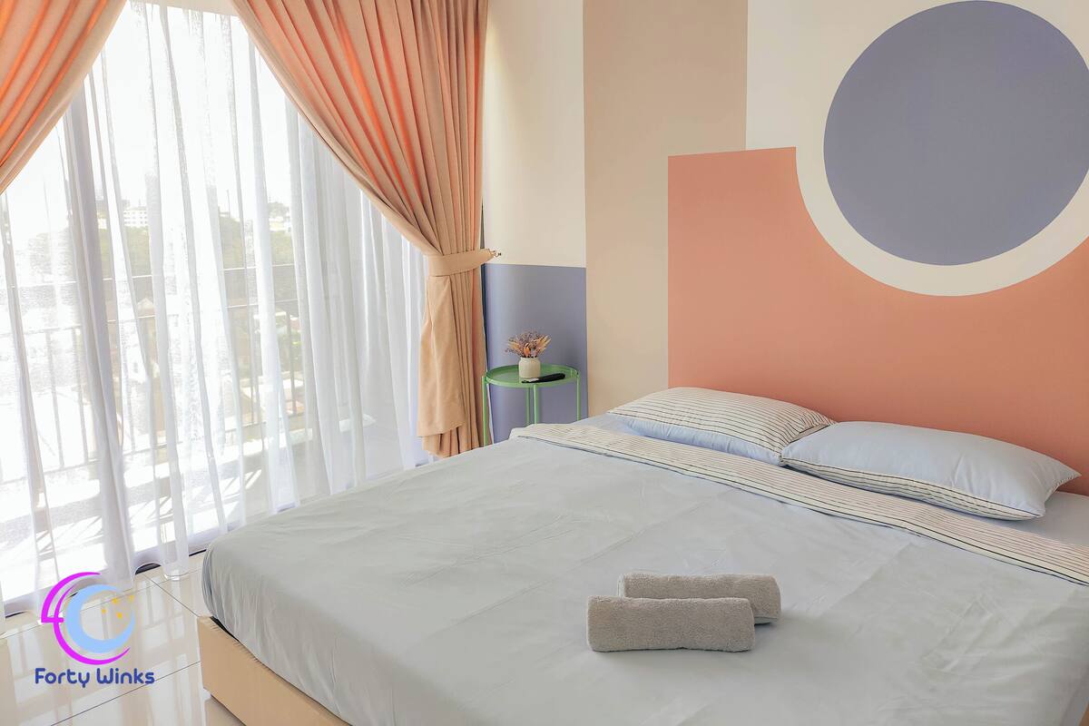 A hotel room with a bed, pillows, a bedside table with a vase, and a view of the outside.