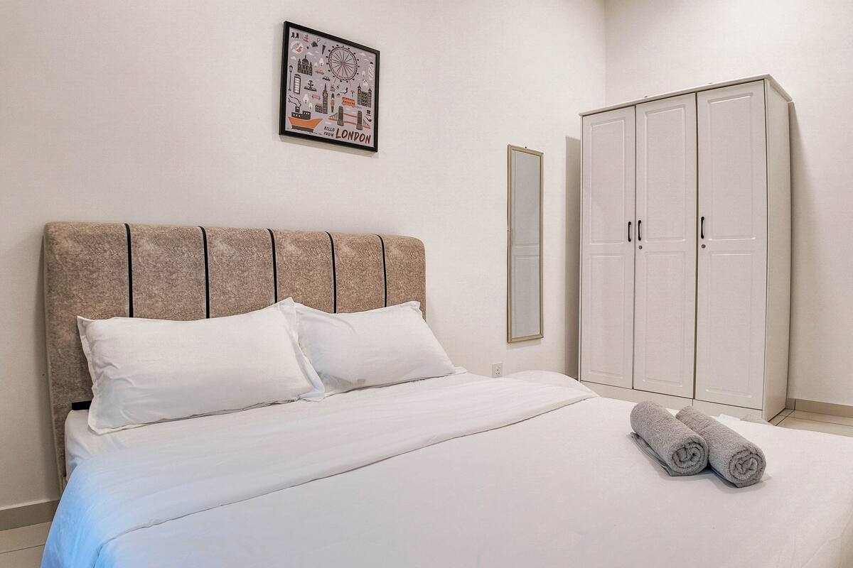 A neatly made bed with white linens, a headboard with a textured pattern, a framed picture on the wall, and a large wardrobe.