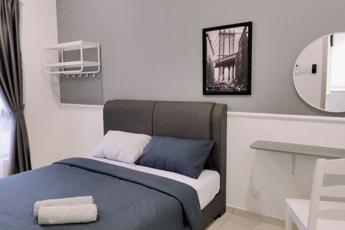 Un dormitorio moderno con una cama gris, almohadas blancas y azules, un cabecero gris, una mesita de noche blanca, un espejo redondo, un cuadro con una imagen de un puente y una toalla blanca sobre la cama.