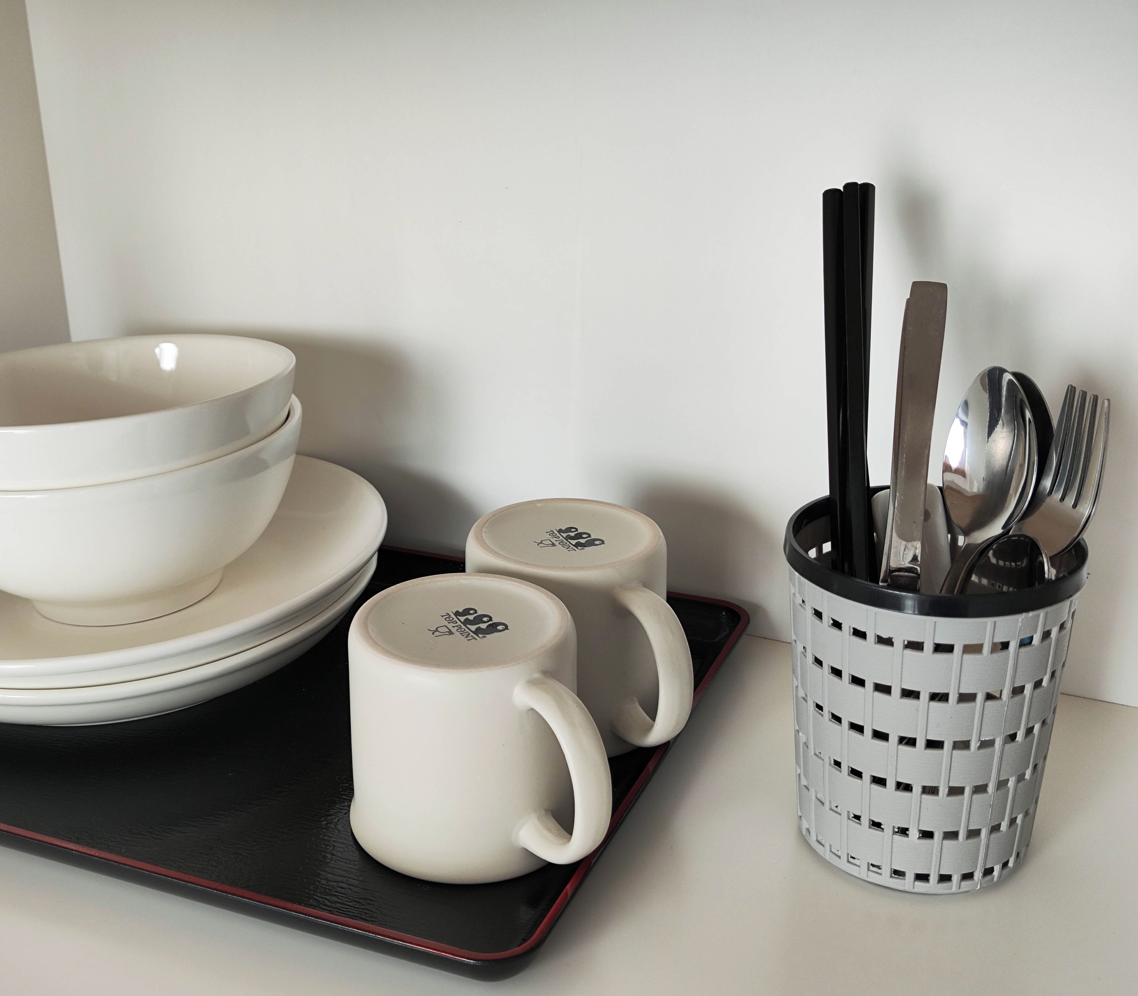A set of white bowls and mugs on a tray, with a utensil holder containing spoons and forks.