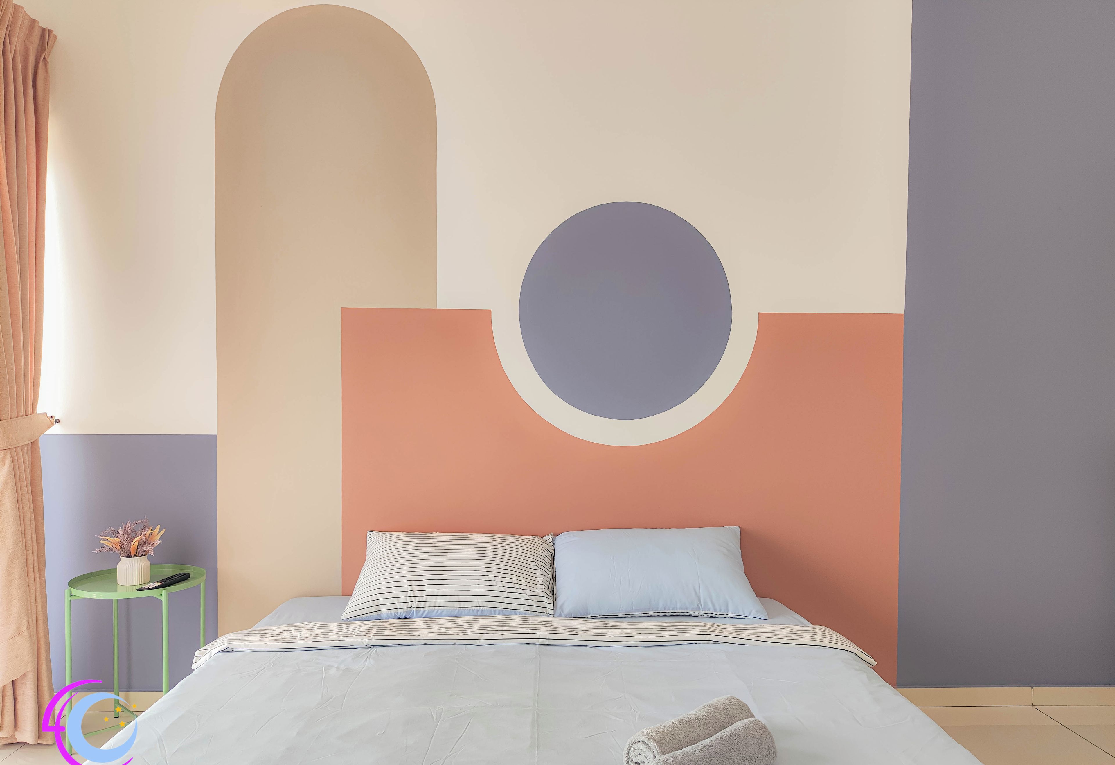 A bedroom with a bed, pillows, a rolled towel, a small table with a vase, and a wall with geometric shapes.