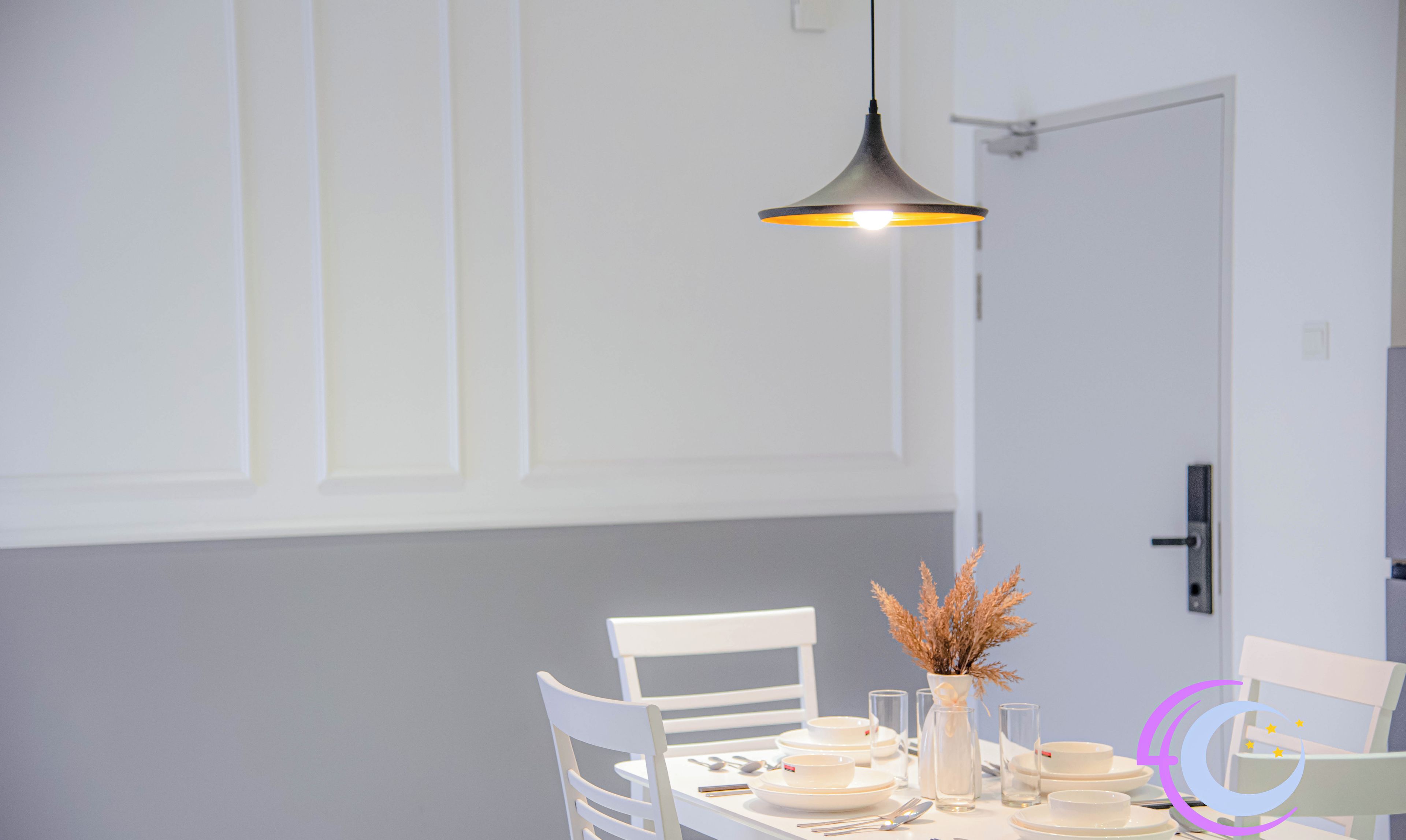 Comedor con una mesa blanca dispuesta para cuatro, incluyendo platos, cubiertos y un jarrón con flores secas. Una lámpara colgante ilumina la mesa.
