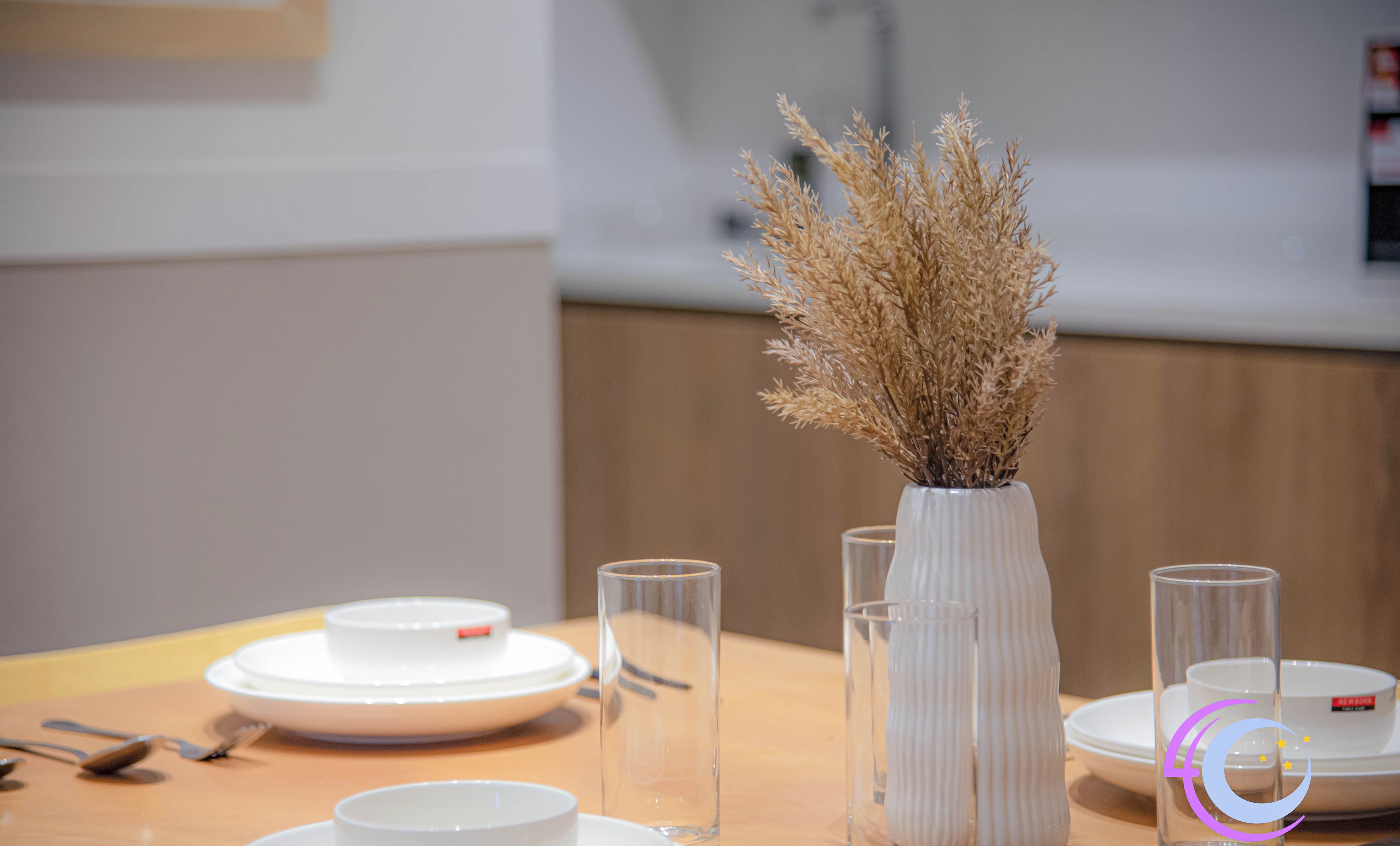 A dining table set with white plates, bowls, and glasses, and a vase with dried plants.