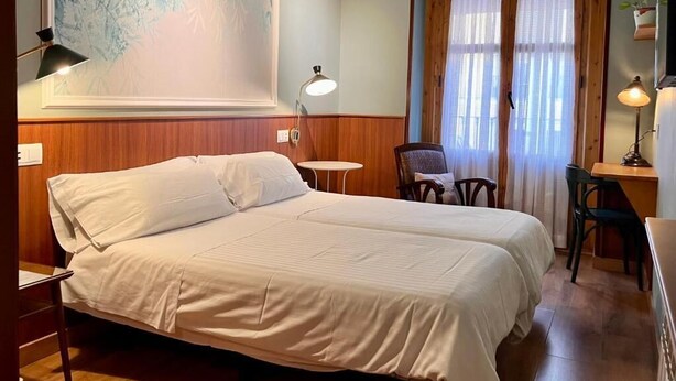 Ein Doppelbett mit weißer Bettwäsche, ein kleiner runder Tisch, zwei Stühle, ein Schreibtisch und ein großes Fenster mit Vorhängen.