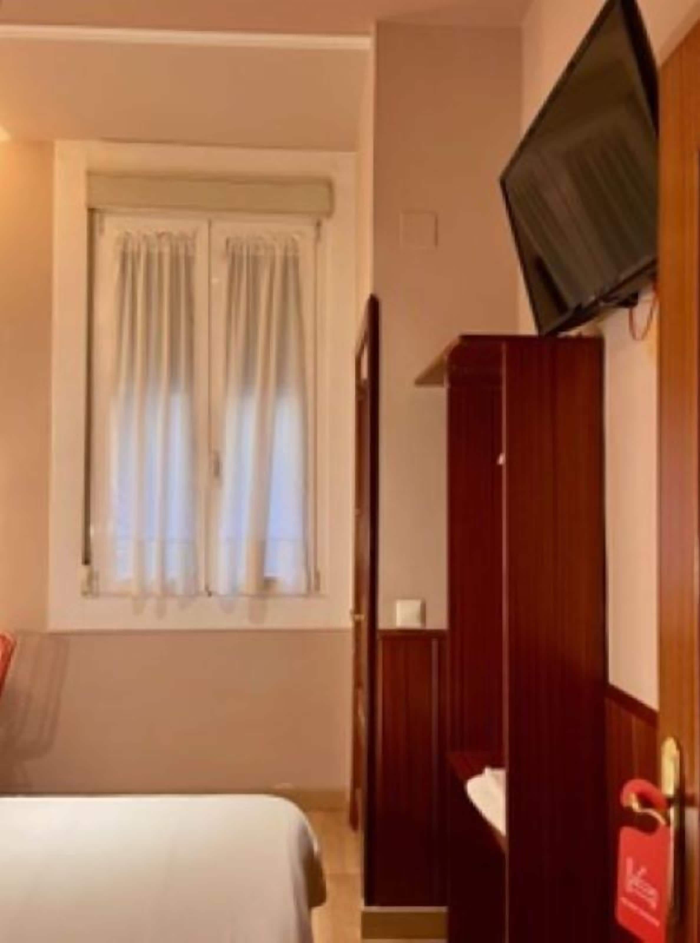Een hotelkamer met een bed, een aan de muur bevestigde televisie, een raam met gordijnen en een houten garderobekast.