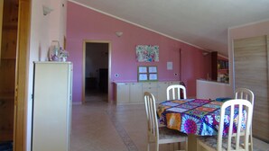 A dining area with a table set for four, a refrigerator, and a kitchen area in the background. - Casa Da Pina B&B (Cabras)