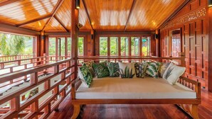 A wooden deck with a cushioned seating area, surrounded by wooden railings, and large windows offering a view of greenery. - 6BR Villa Huge Pool & Mountain View V89 (Rawai)