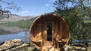 Una cabaña de madera con dos bancos de madera y una puerta, ubicada junto a un lago con montañas al fondo.