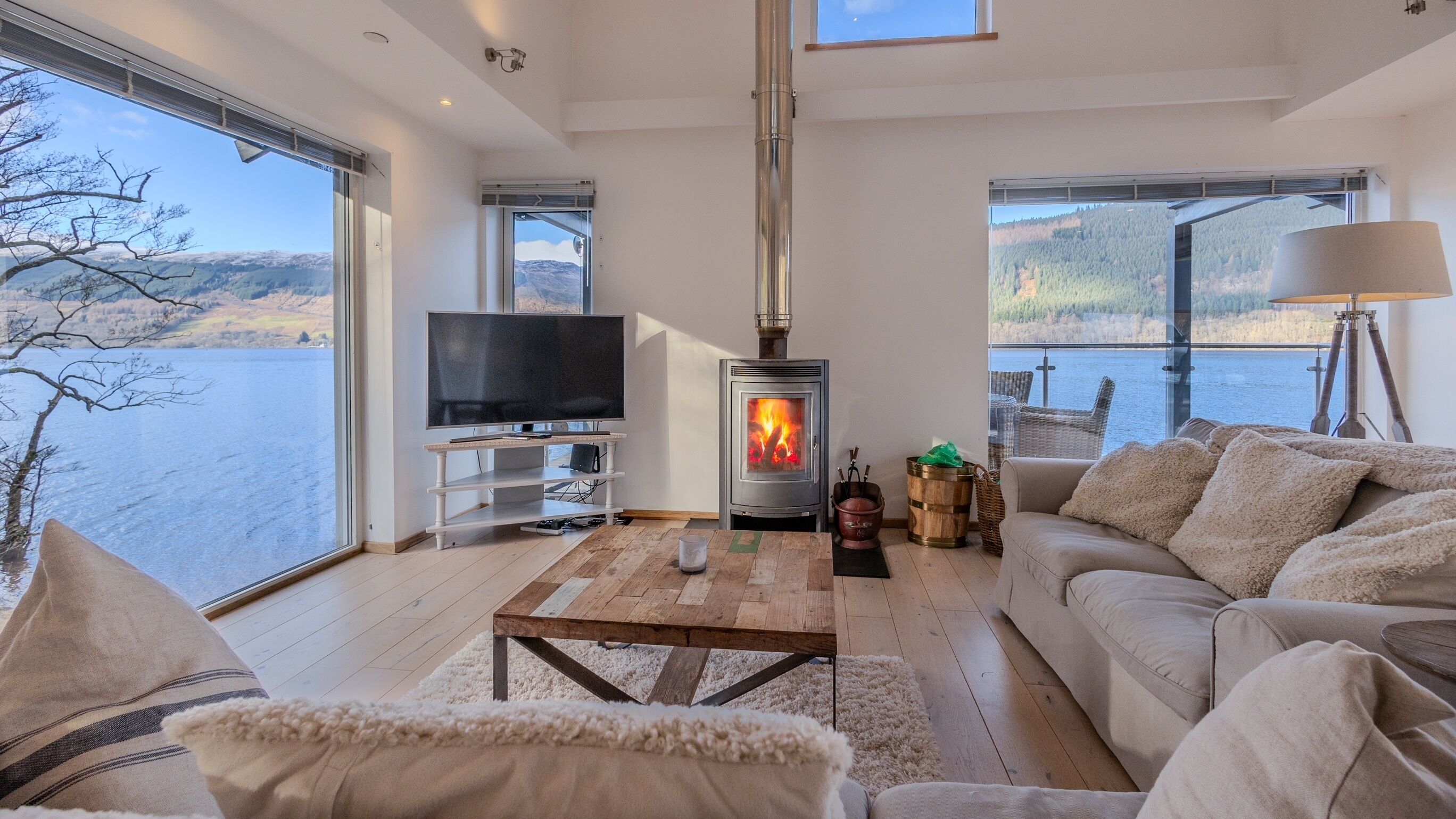 A modern living room with a large window overlooking a lake, a fireplace, a television, and a cozy seating area.