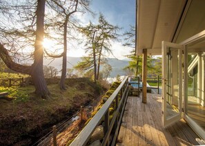 Una terraza de madera con un ventanal de vidrio que da a un área boscosa, con vistas a montañas y una masa de agua.