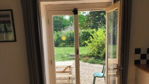 Ein Durchgang zu einer Terrasse im Freien mit einem Holztisch und Stühlen, umgeben von Grünpflanzen und einem an der Wand befestigten Feuerlöscher.