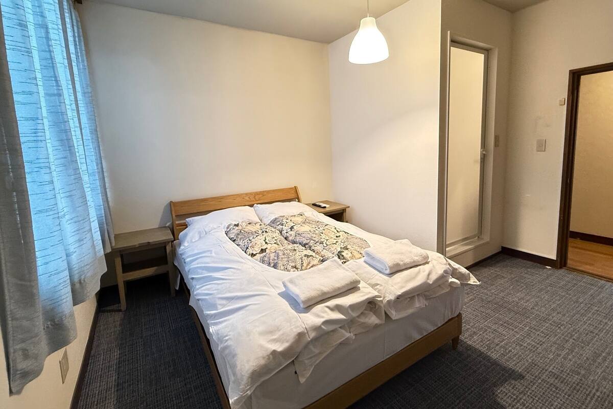 A neatly made bed with a floral comforter and white linens, a bedside table with a remote control, and a hanging pendant light.