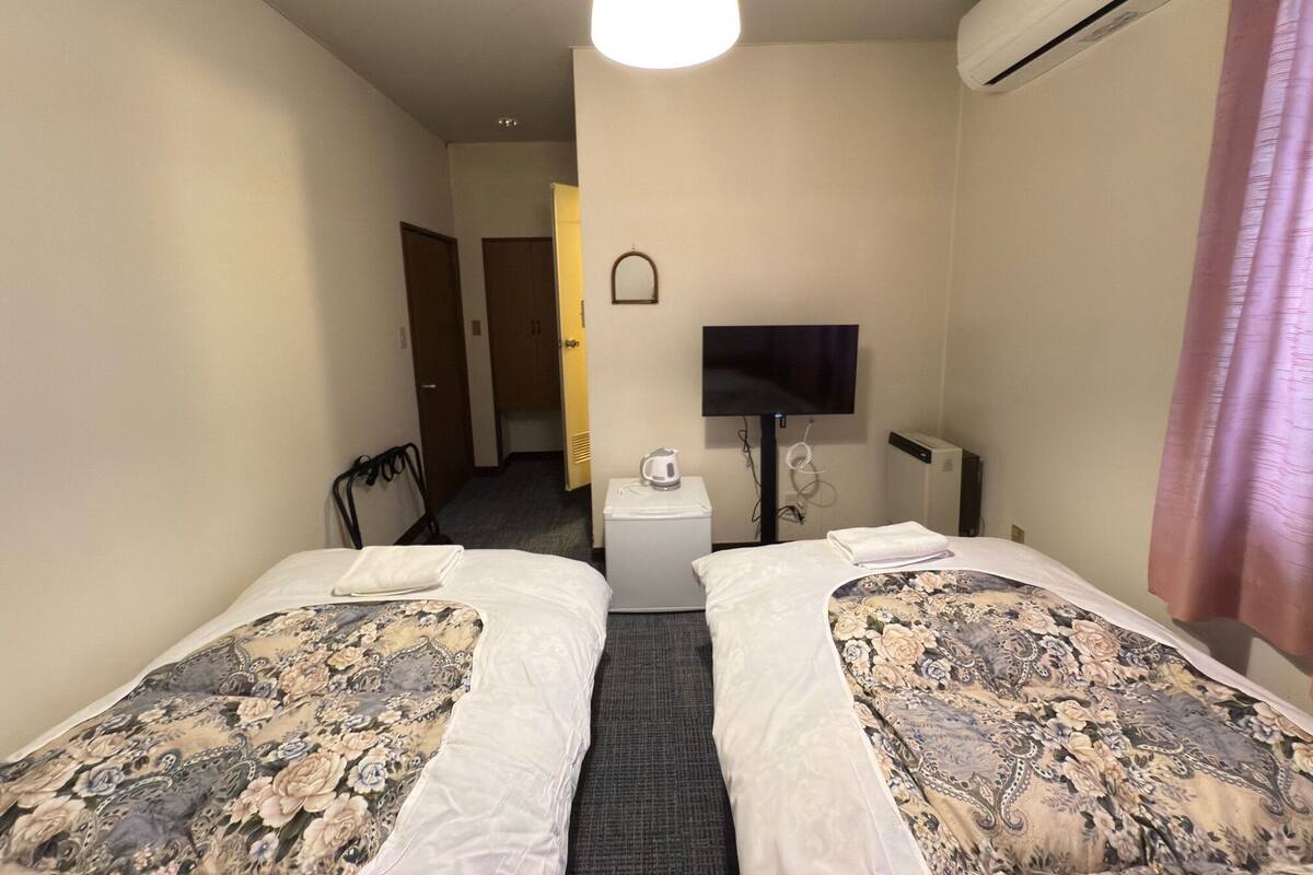 A hotel room with two beds, a television, an air conditioning unit, and a small table with a cup.