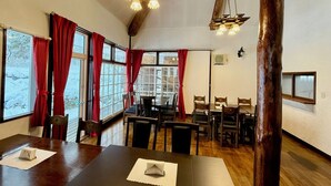 Un espace repas avec des tables et des chaises en bois, de grandes fenêtres ornées de rideaux rouges et un plafond en bois agrémenté de luminaires suspendus.
