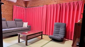 Une pièce avec un mur en bois, un canapé gris, un rideau rouge, un sol recouvert de tatamis et un téléviseur.