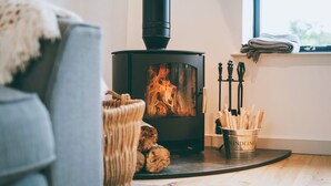 Un accogliente soggiorno con stufa a legna, un divano grigio, un cesto intrecciato con legna e un secchio in metallo con l'etichetta 'KINDLING' su un pavimento in legno.