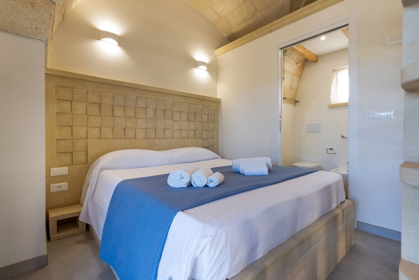Una habitación de hotel con una cama grande, un cabecero de madera y un baño visible a través de una puerta abierta.