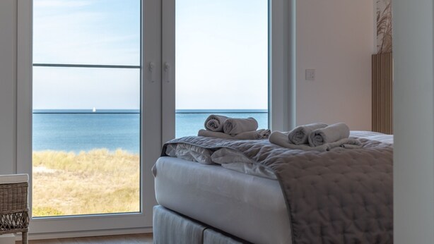 A bedroom with a large window overlooking the sea, a bed with rolled towels, and a wicker chair. - Wonderfully modern vacation apartment with a direct view of the Baltic Sea (Graal-Müritz)