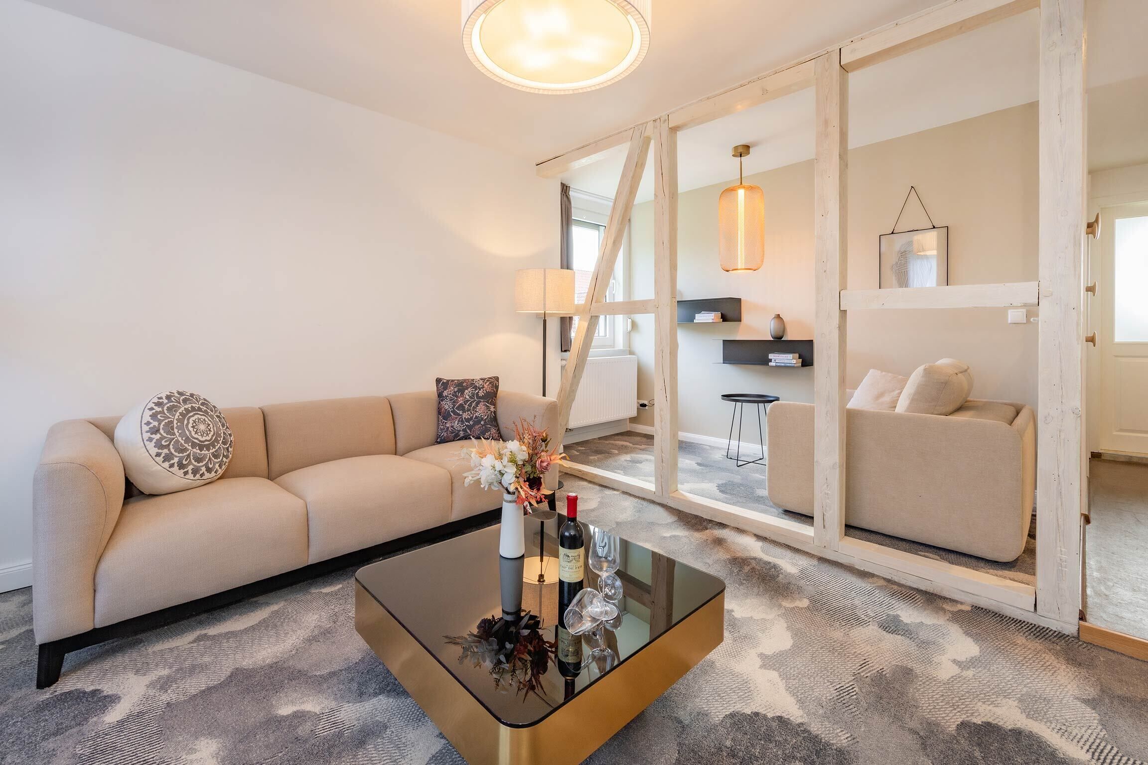 A modern living room with a beige sofa, a glass coffee table with a vase and wine glasses, and a view through a glass door to another room.