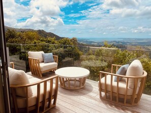 A balcony with wooden furniture, a glass barrier, and a view of a hilly landscape. - Matakana View- Treetop retreat in Matakana (Whangaripo)