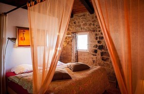 A bedroom with a stone wall, a bed with floral bedding, and a window with curtains. - Le Mas Bleu (Rosières)