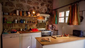A rustic kitchen with a stone wall, wooden shelves, a stove, and a window with colorful curtains. - Le Mas Bleu (Rosières)