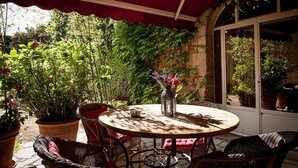 A round table with wicker chairs under a red awning, surrounded by potted plants and a stone wall. - Le Mas Bleu (Rosières)