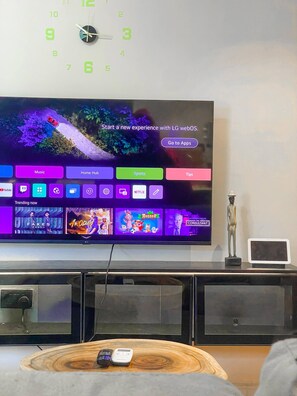 A living room with a flat-screen TV mounted on the wall displaying a home screen with various app icons and a clock above it. - Peaceful Abuja Retreat - 1 Bedroom Private Apartment (Abuja)