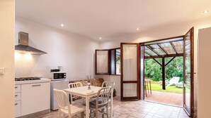 Cocina compacta con armarios blancos, cocina a gas y microondas. Hay una mesa de comedor con cuatro sillas y un banco estampado. Una puerta de cristal da a un patio exterior con vistas a zonas verdes.